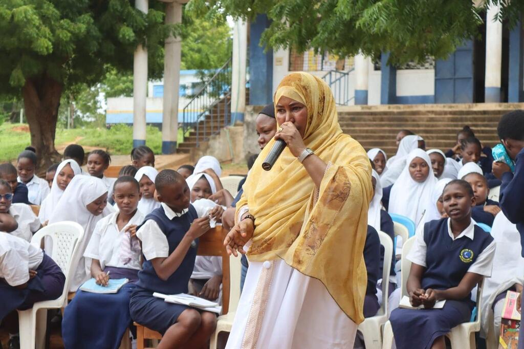 Mwanamke katika hijab ya manjano akizungumza kupitia kipaza sauti kwa kundi la wanafunzi walioketi kwenye viti katika sherehe ya shule ya nje huko Tana River, Kenya, kama sehemu ya Mpango wa Jamii Nzuri.