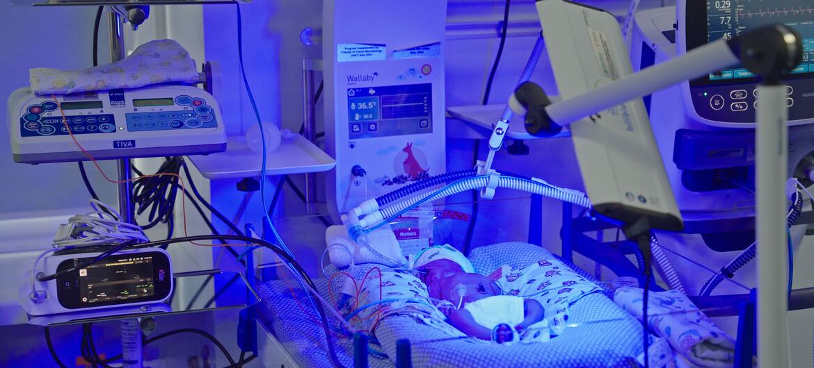 Новорожденный лежит в инкубаторе в родильной больнице в Украине, окруженный медицинским оборудованием, таким как мониторы и системы кислорода. Сцена подчеркивает критические потребности в здравоохранении на фоне продолжающегося конфликта и повреждения инфраструктуры.
