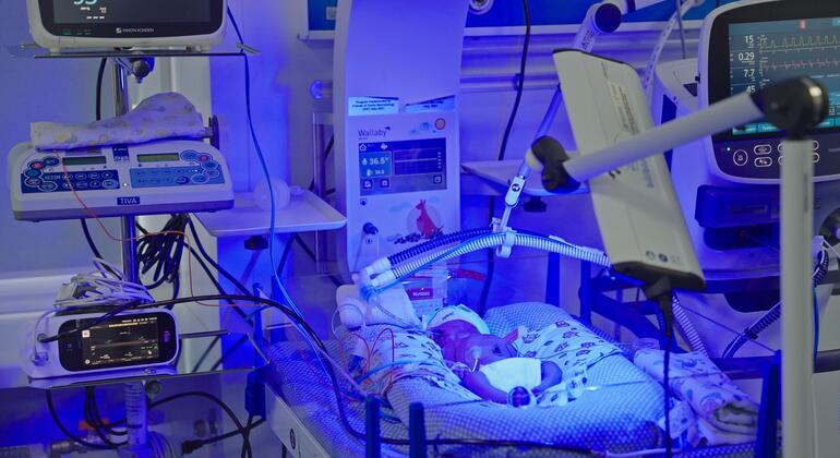 Um recém-nascido está deitado em uma incubadora em um hospital de maternidade na Ucrânia, cercado por equipamentos médicos, como monitores e sistemas de oxigênio. A cena destaca as necessidades críticas de cuidados de saúde em meio ao conflito em curso e danos à infraestrutura.