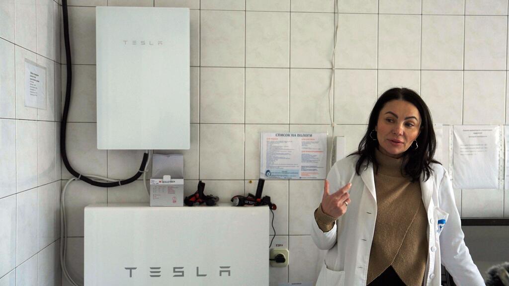 Медицинский работник в белом пальто стоит рядом с энергоблоком Tesla в украинской родильной больнице, подчеркивая усилия по поддержанию критической помощи во время отключения электроэнергии.