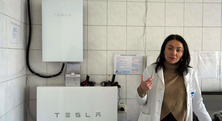 Медицинский работник в белом пальто стоит рядом с энергоблоком Tesla в украинской родильной больнице, подчеркивая усилия по поддержанию критической помощи во время отключения электроэнергии.