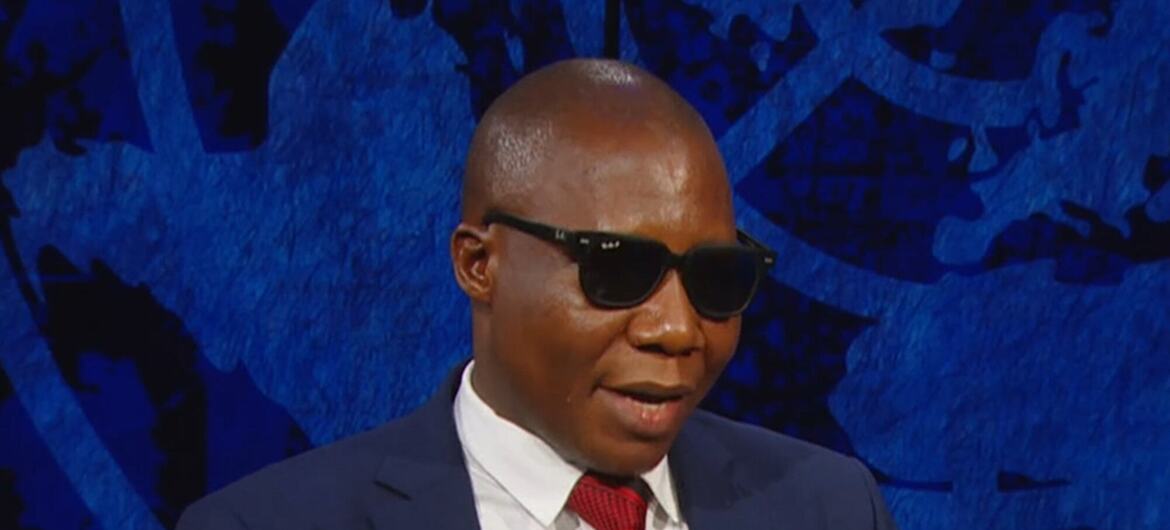 Mwanadiplomasia wa Tanzania amevaa miwani, suti, na tai, akifanya ishara wakati akizungumza katika hafla ya Umoja wa Mataifa iliyozingatia SDGs na CSocD64.