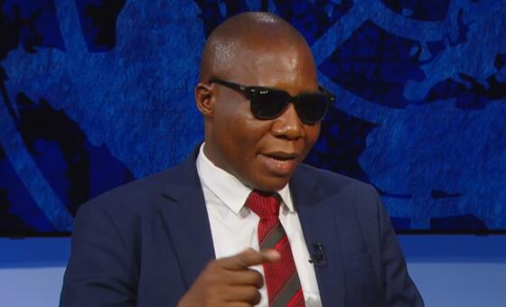 Mwanadiplomasia wa Tanzania amevaa miwani, suti, na tai, akifanya ishara wakati akizungumza katika hafla ya Umoja wa Mataifa iliyozingatia SDGs na CSocD64.