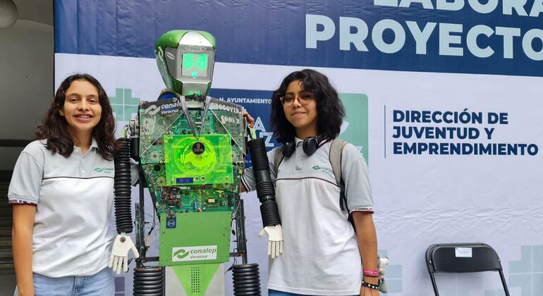 Dos jóvenes mexicanas están junto a un robot que crearon, mostrando su proyecto de ciencia e ingeniería en un evento para jóvenes y emprendedores.