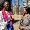 Wanawake weusi wawili wenye mavazi ya kitaalamu wakizungumza katika bustani yenye mandhari ya jiji nyuma.