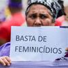 Uma mulher brasileira em um protesto segura um cartaz que diz 'BASTA DE FEMINICÍDIOS' (Basta de Feminicídios) e carrega um monte de plantas verdes.