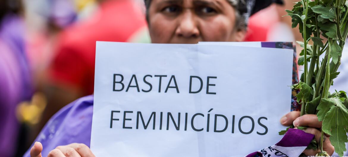 Uma mulher brasileira em um protesto segura um cartaz que diz 'BASTA DE FEMINICÍDIOS' (Basta de Feminicídios) e carrega um monte de plantas verdes.