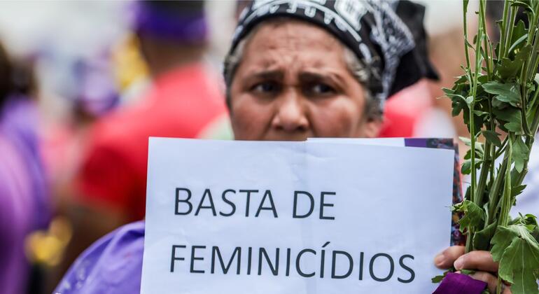 Uma mulher brasileira em um protesto segura um cartaz que diz 'BASTA DE FEMINICÍDIOS' (Basta de Feminicídios) e carrega um monte de plantas verdes.