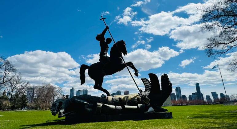 घोड़े पर सवार सेंट जॉर्ज की एक काली धातु की प्रतिमा, जो एक ड्रैगन को मार रही है, एक घास से ढके पार्क में स्थित है, जिसके पृष्ठभूमि में बादलों वाले नीले आकाश के नीचे न्यूयॉर्क शहर की आकाशरेखा दिखाई दे रही है।
