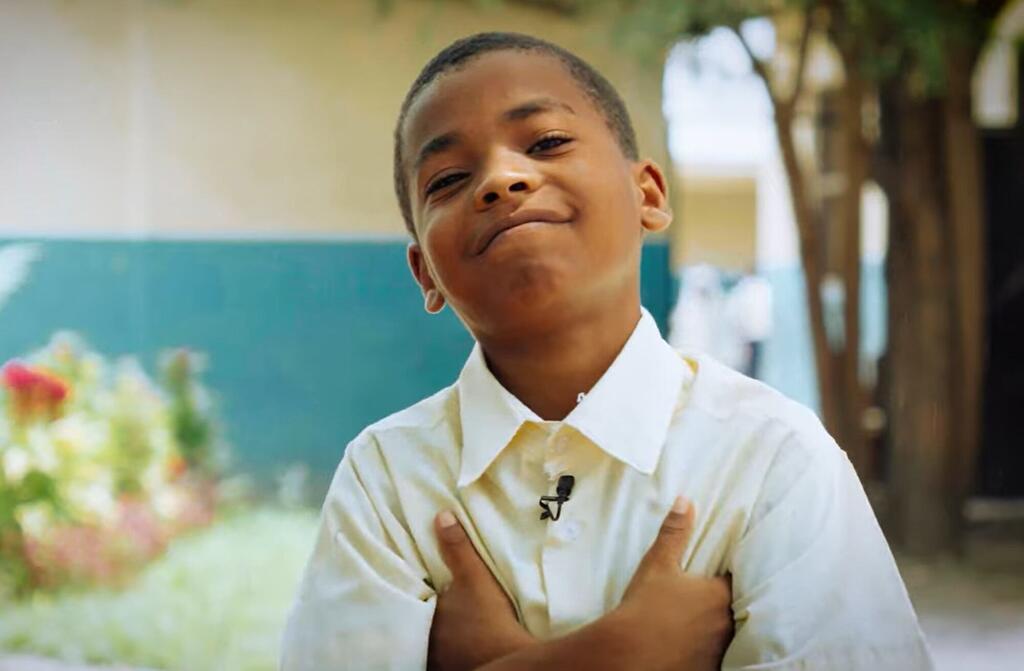 Mvulana mdogo wa Kiafrika aliyevaa sare nyeupe ya shule anatabasamu mbele ya kamera akiwa na mikono kwenye kifua chake, akiwa amevaa kipaza sauti kidogo. Yeye yuko nje huko Zanzibar, Tanzania, kama sehemu ya mpango wa UNICEF wa uhifadhi wa mazingira na elimu.