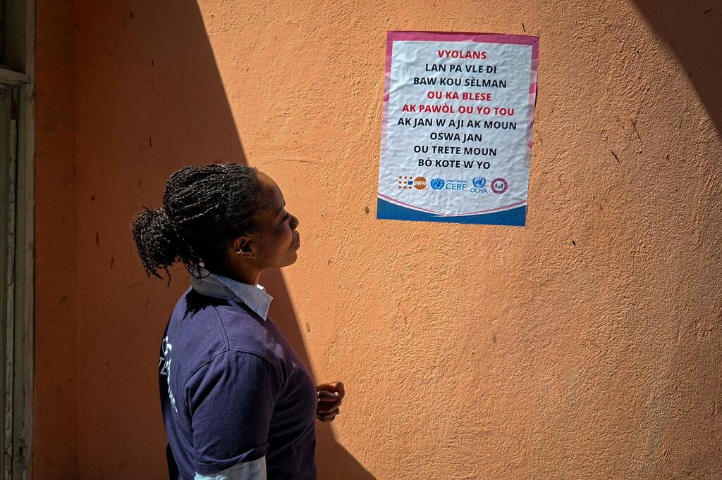 A Black woman with braided hair stands in profile looking at a poster on an orange wall in Port-au-Prince, Haiti. The poster is a gender-based violence awareness campaign with logos from UN agencies including UNOCHA, CERF, and UNFPA.