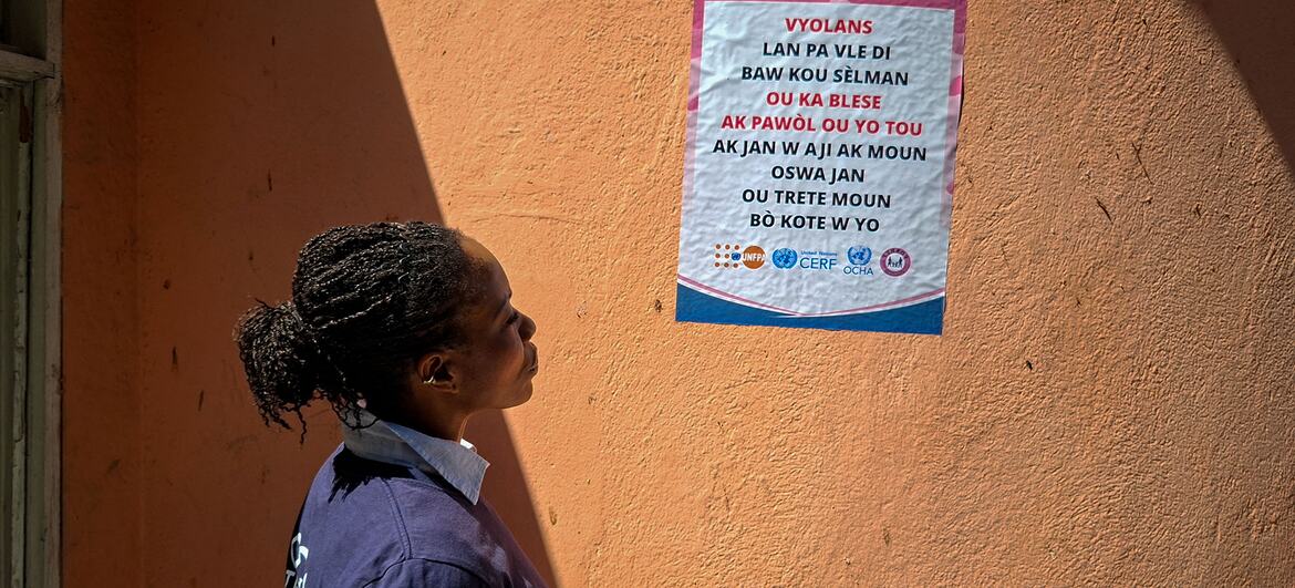 Une femme noire aux cheveux tressés se tient de profil en regardant une affiche sur un mur orange à Port-au-Prince, en Haïti. L'affiche est une campagne de sensibilisation à la violence basée sur le genre, avec les logos d'agences des Nations Unies, notamment l'UNOCHA, le CERF et l'UNFPA.