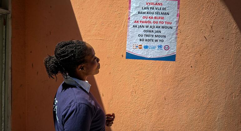 A Black woman with braided hair stands in profile looking at a poster on an orange wall in Port-au-Prince, Haiti. The poster is a gender-based violence awareness campaign with logos from UN agencies including UNOCHA, CERF, and UNFPA.