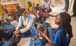 Edem Wosornu, Director of OCHA Crisis Response Division, sits on a blue mat with displaced women and children at a Community Resource Centre in Delmas, Haiti, supported by IOM.