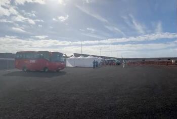 Red bus and white tents set up outdoors on gravel ground under a blue sky, with people in protective gear near the tents.