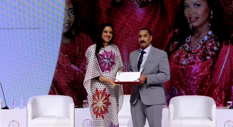 رجل يرتدي بدلة رمادية يقدم شهادة جائزة لامرأة ترتدي ثوباً مرسومًا في حفل توزيع جوائز هيوم في المنامة، البحرين.