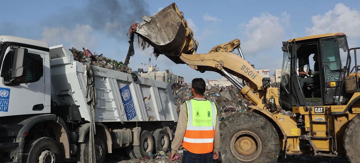 عامل يرتدي سترة أمان يشاهد محفرة صفراء تحمل القمامة في شاحنة قمامة تحمل العلامة التجارية لبرنامج الأمم المتحدة الإنمائي في منطقة النفايات في سوق الفارس في مدينة غزة.