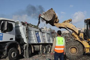 عامل يرتدي سترة أمان يشاهد محفرة صفراء تحمل القمامة في شاحنة قمامة تحمل العلامة التجارية لبرنامج الأمم المتحدة الإنمائي في منطقة النفايات في سوق الفارس في مدينة غزة.