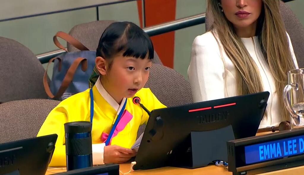 A young girl named Emma Lee, wearing a yellow traditional outfit, speaks into a microphone at a UN conference, with a nameplate in front of her.