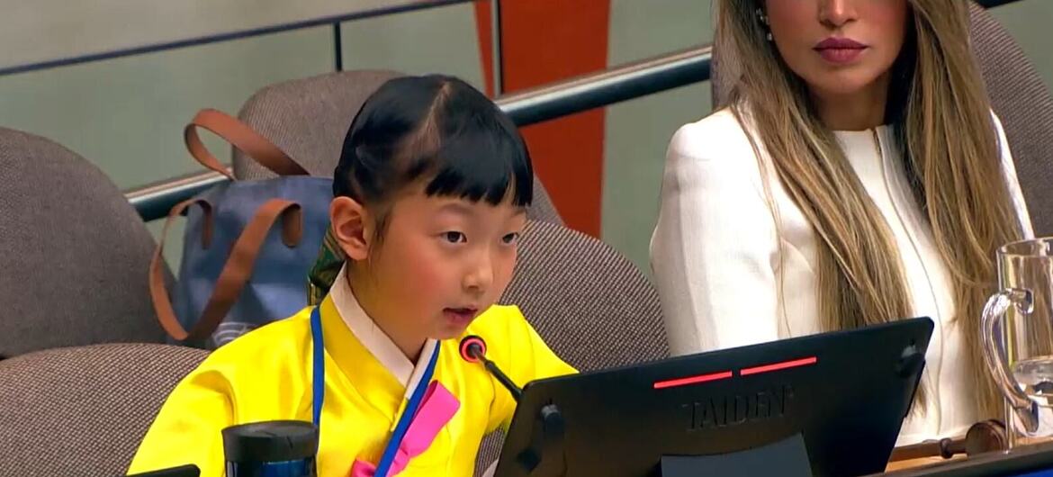 A young girl named Emma Lee, wearing a yellow traditional outfit, speaks into a microphone at a UN conference, with a nameplate in front of her.