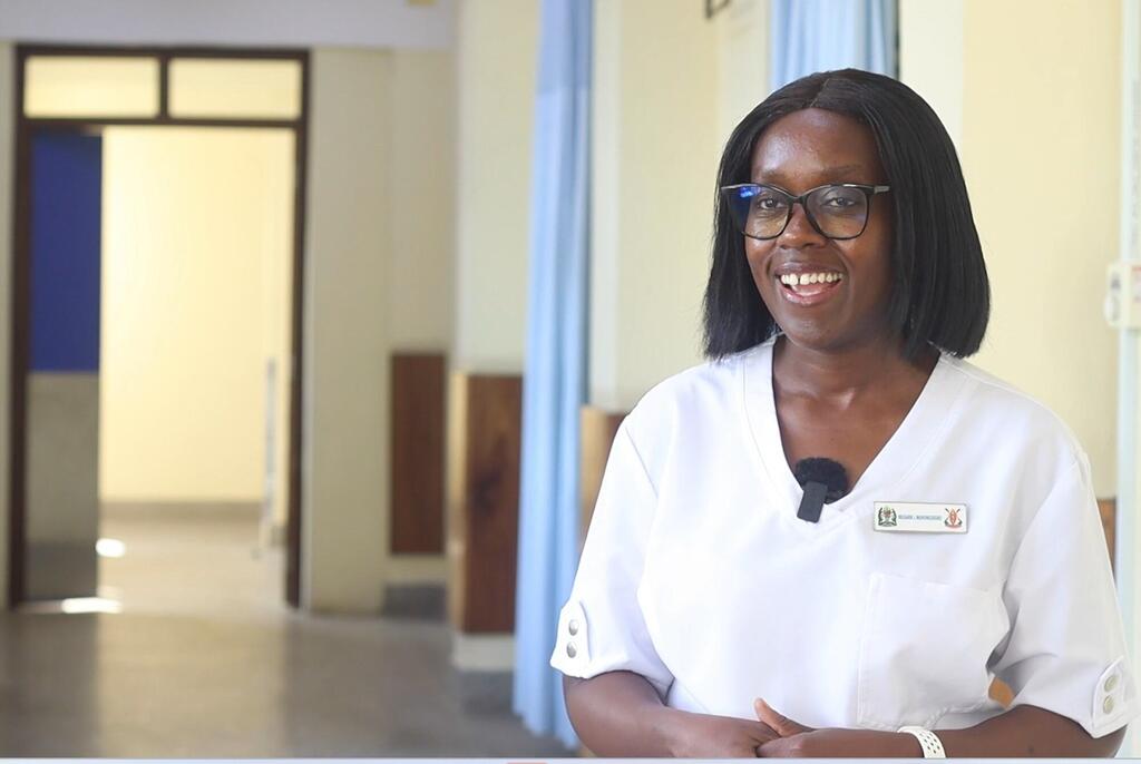 Mwanamke mwenye tabasamu kutoka Tanzania akiwa amevaa kanzu nyeupe ya daktari mwenye lebo ya jina, amesimama ndani ya nyumba katika mazingira ya kitaalamu.