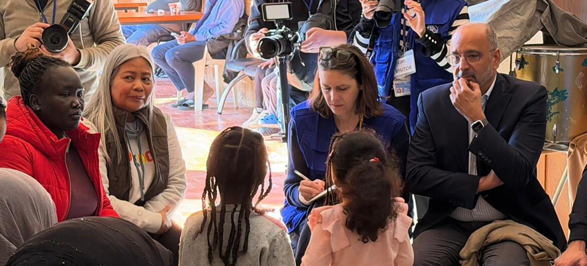 يجلس عمران رضا، منسق الأمم المتحدة المقيم ومنسق المساعدات الإنسانية في لبنان، ويستمع إلى النازحين الذين اضطروا إلى الإجلاء إلى أماكن آمنة. يحيط به مجموعة من النساء والأطفال، ويمكن رؤية أفراد وسائل الإعلام والكاميرات في الخلفية.