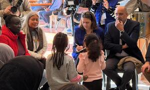 Imran Riza, UN Resident Coordinator and Humanitarian Coordinator in Lebanon, is seated and listening to displaced people who had to evacuate to safety. He is surrounded by a group of women and children, with media personnel and cameras visible in the background.