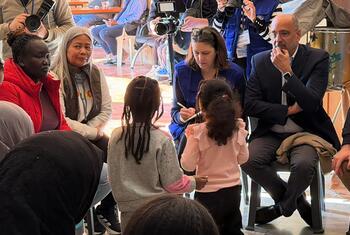 يجلس عمران رضا، منسق الأمم المتحدة المقيم ومنسق المساعدات الإنسانية في لبنان، ويستمع إلى النازحين الذين اضطروا إلى الإجلاء إلى أماكن آمنة. يحيط به مجموعة من النساء والأطفال، ويمكن رؤية أفراد وسائل الإعلام والكاميرات في الخلفية.