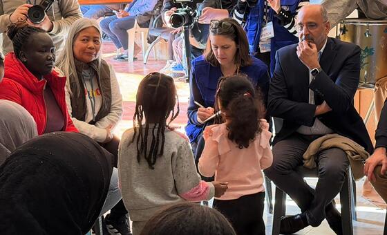 Imran Riza, UN Resident Coordinator and Humanitarian Coordinator in Lebanon, is seated and listening to displaced people who had to evacuate to safety. He is surrounded by a group of women and children, with media personnel and cameras visible in the background.