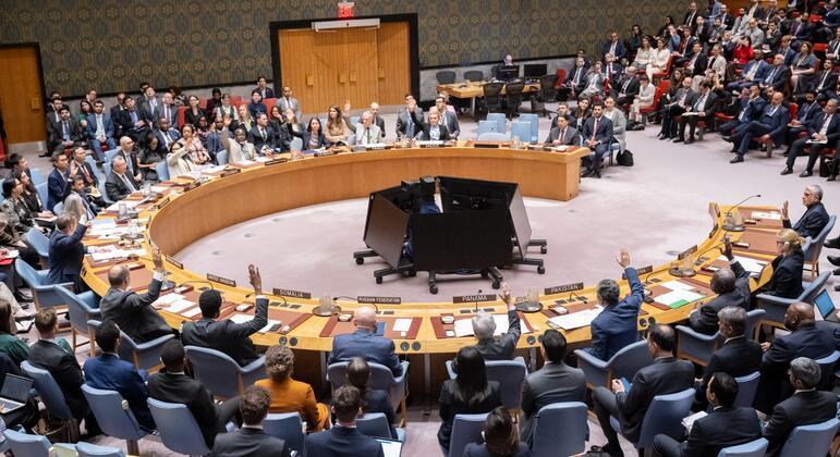 A wide shot of the United Nations Security Council chamber showing delegates seated around a large circular table, with several members raising their hands to vote. The room is filled with attendees observing the proceedings.