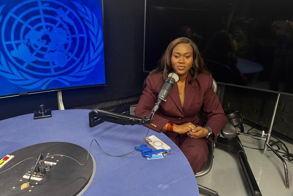 Mwanamke katika suti ya maroon ameketi kwenye kipaza sauti katika studio ya redio akiwa na nembo ya Umoja wa Mataifa kwenye skrini nyuma yake.