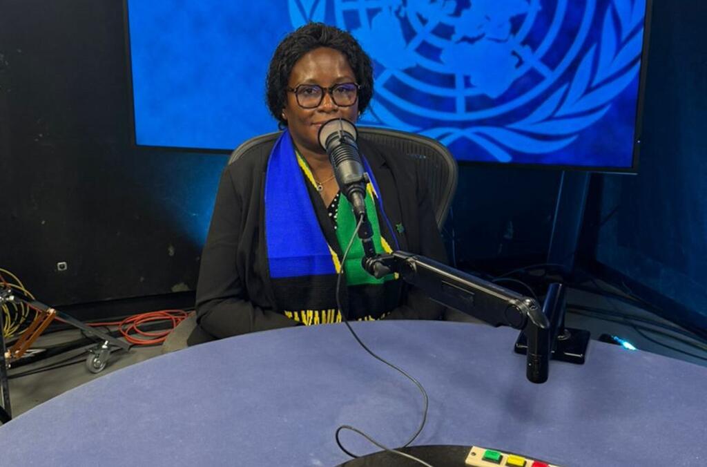 Mwanamke mweusi aliyevaa miwani na shati ameketi mezani akizungumza kwenye kipaza sauti na ishara ya Umoja wa Mataifa nyuma.