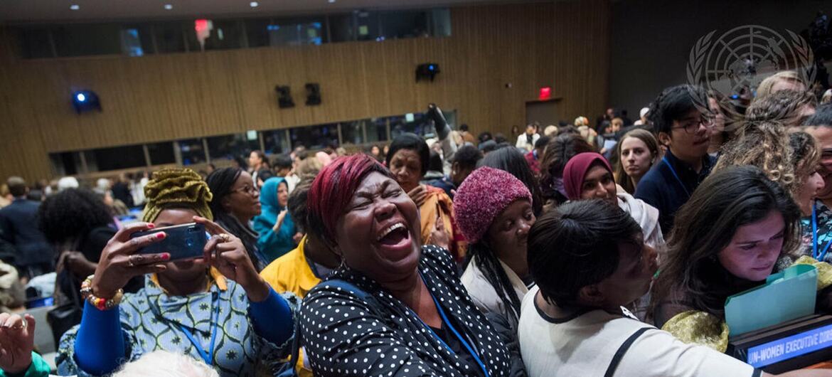 Un groupe diversifié de femmes dans une salle de conférence, avec une femme noire en premier plan en train de rire et d'applaudir avec enthousiasme tout en serrant la main à quelqu'un en dehors de la caméra.
