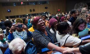 Un groupe diversifié de femmes dans une salle de conférence, avec une femme noire en premier plan en train de rire et d'applaudir avec enthousiasme tout en serrant la main à quelqu'un en dehors de la caméra.