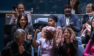 Kikundi tofauti cha wanawake wa makabila mbalimbali wameketi katika ukumbi, wakipiga makofi na kutabasamu kwenye mkutano. Nembo ya Umoja wa Mataifa inaonekana nyuma.