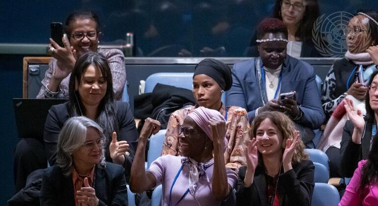Kikundi tofauti cha wanawake wa makabila mbalimbali wameketi katika ukumbi, wakipiga makofi na kutabasamu kwenye mkutano. Nembo ya Umoja wa Mataifa inaonekana nyuma.