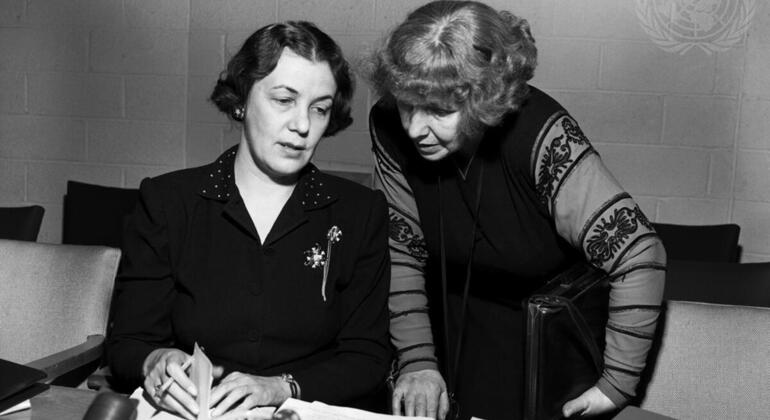 Picha nyeusi na nyeupe ya wanawake wawili, waliotambuliwa kama Dorothy Hodgkin na Eleanor Roosevelt, wakitazama nyaraka kwenye meza ya mkutano na nembo ya Umoja wa Mataifa ikionekana kwenye ukuta nyuma yao.