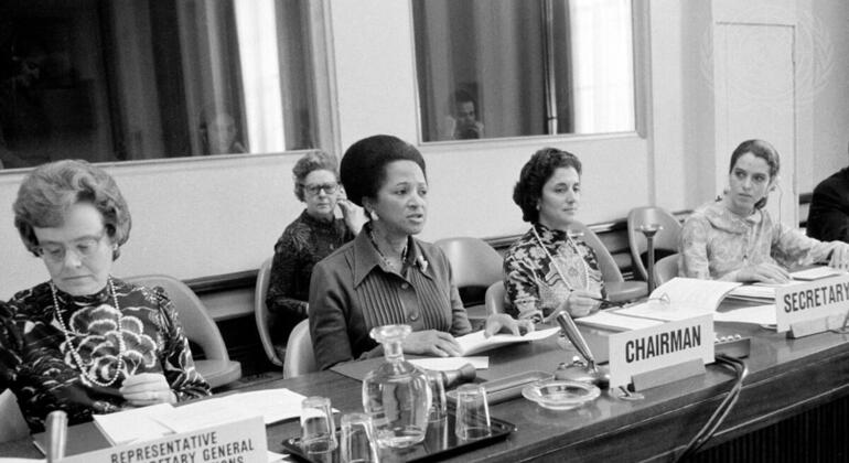 Picha nyeusi na nyeupe ya wanawake kwenye meza rasmi ya mkutano huko Geneva, ikiwa ni pamoja na Eugenia Charles, pamoja na ishara za 'Wakilishi wa Katibu Mkuu wa Umoja wa Mataifa' na 'Rais'.