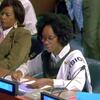 Duas mulheres africanas representando Moçambique em uma conferência internacional. A mulher em primeiro plano está escrevendo enquanto a mulher em segundo plano está assistindo. Uma placa azul com "MOZAMBIQUE" é visível na mesa.