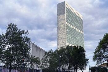The United Nations Secretariat Building in New York, a tall modernist tower with a concrete and glass facade, viewed from street level among trees under a cloudy sky.
