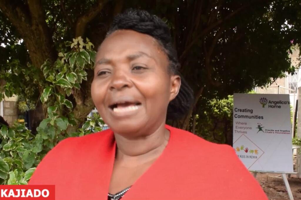 Mwanamke mweusi aliyevaa koti jekundu anaongea wakati wa mahojiano ya nje mbele ya alama ya Angelica's Home huko Kajiado, Kenya.