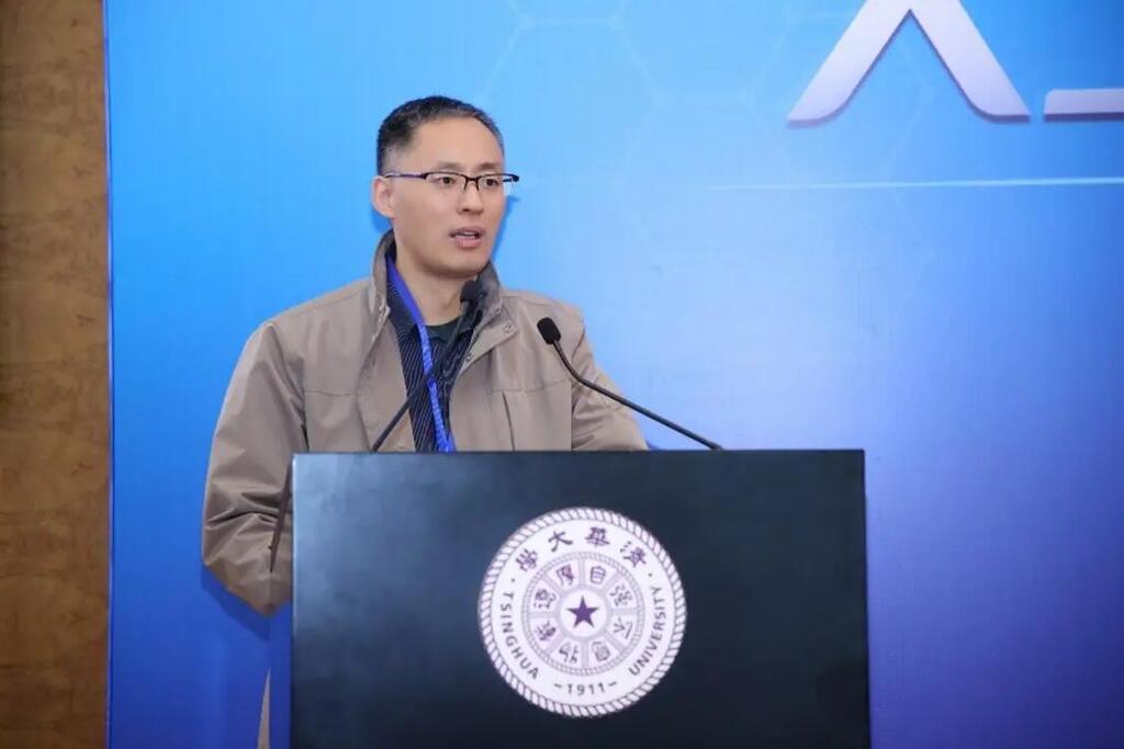 Professor Shaojing Sun speaking at a podium with the Tsinghua University logo, wearing glasses and a beige jacket.