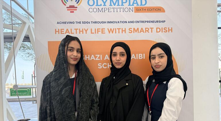ثلاث طلابات من مدرسة غازي أ. العيبي، تظهر للصورة في المسابقة الأولمبية WEIF2026 في البحرين، وقوفهن أمام لافتة تعزز "الحياة الصحية مع الأطباق الذكية".