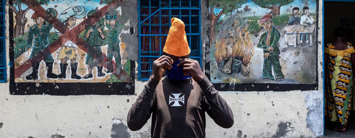 A young person adjusts a face mask while standing in front of murals depicting de-mobilization at a UNICEF-supported transit center for children in Goma, DRC.