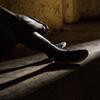 Um menino sentado sozinho em uma sala pouco iluminada, com as pernas levantadas, iluminadas por um pedaço de luz no chão de concreto, refletindo seu trauma de ser criança soldado na República Democrática do Congo.
