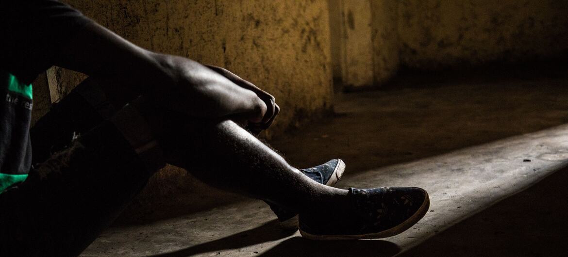 Un jeune garçon est assis seul dans une pièce faiblement éclairée, les jambes tendues, éclairées par une lampe sur le sol en béton, reflétant son traumatisme d'enfant soldat en République démocratique du Congo.