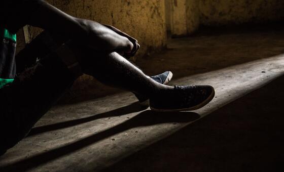 A young boy sits alone in a dimly lit room, his legs drawn up, illuminated by a sliver of light on the concrete floor, reflecting his trauma from being a child soldier in the Democratic Republic of the Congo.