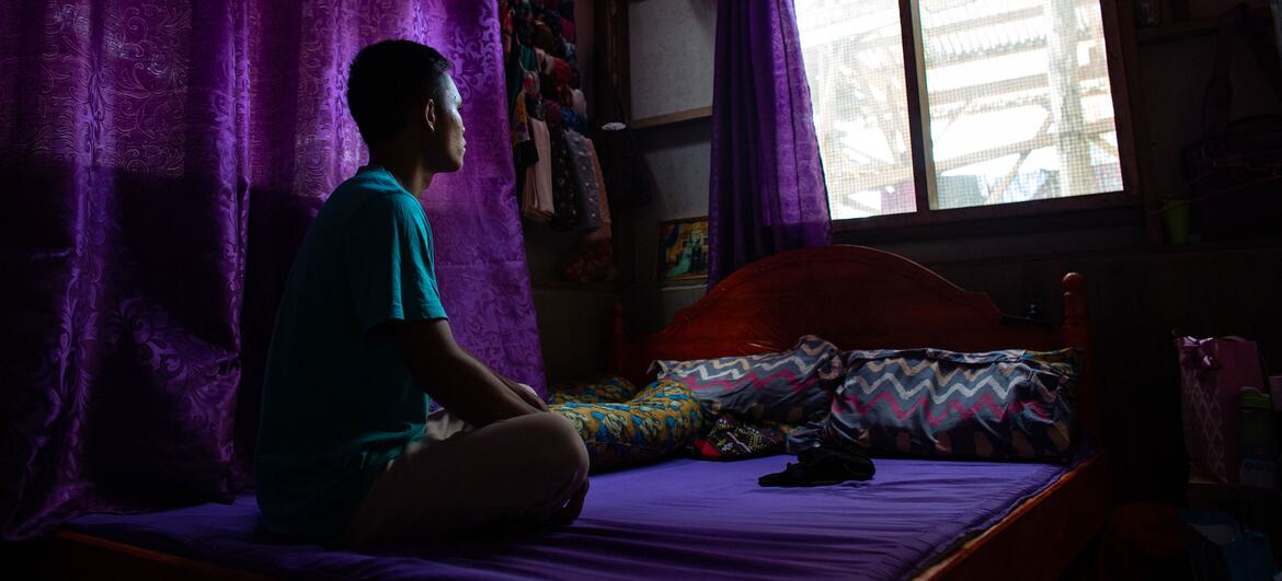 A young boy sits on his bed in a dimly lit room with purple curtains, looking out the window. The image captures a moment of reflection, symbolizing his journey from military service to rejoining education and dreaming of becoming a social worker.