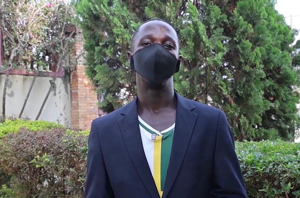 Mwanamume amevaa kinyago cha uso mweusi na suti na kanzu ya kijani, njano, na nyeupe, amesimama nje akiwa na mazingira ya kijani na ukuta wa matofali nyuma.