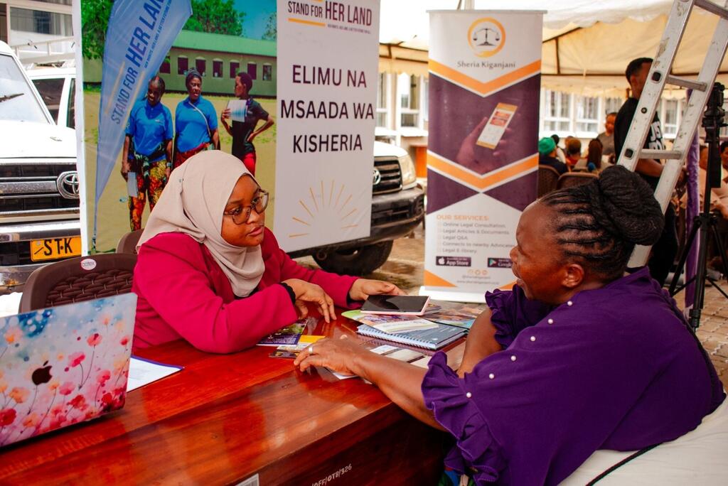 Wanawake wawili wa Kiafrika wanajadiliana kwenye kibanda cha msaada wa kisheria nje ya nyumba, na mabango nyuma yanayoendeleza haki za ardhi na huduma za kisheria kwa Kiswahili.